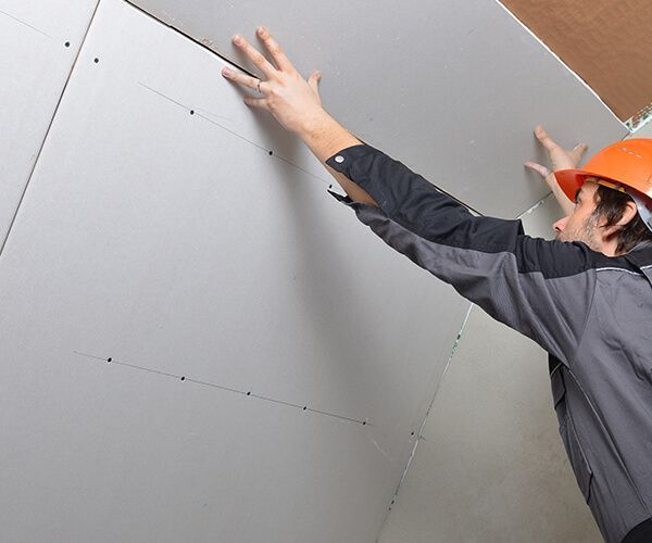 Un ouvrier du bâtiment portant un casque de chantier installe un panneau de plaques de plâtre au plafond.