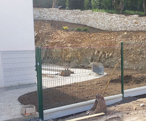 Un panneau de clôture métallique vert est installé devant un mur blanc, avec un paysage rocheux en pente en arrière-plan.