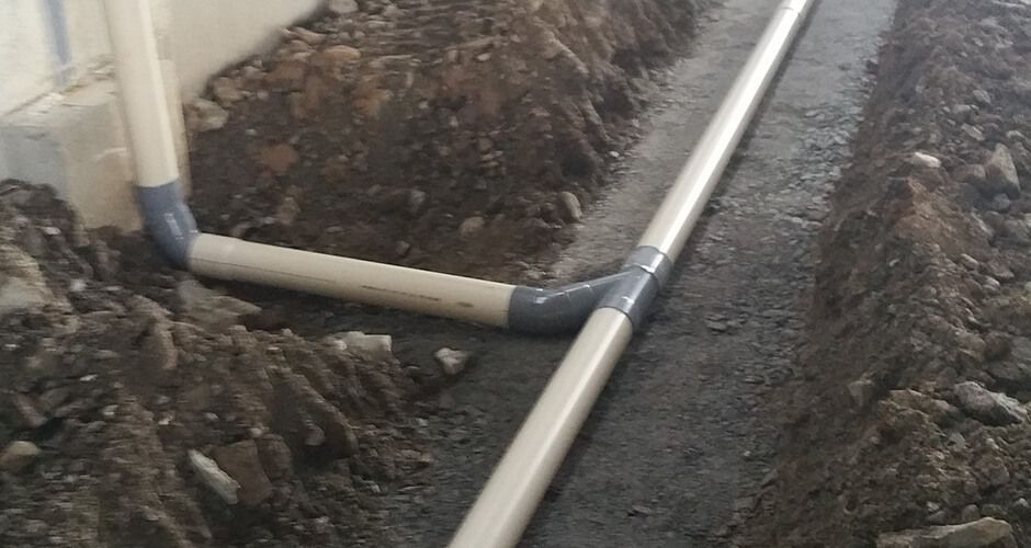 Un ensemble de tuyaux en PVC blanc avec un raccord en Y, installé dans une tranchée de terre le long d'une fondation en béton.