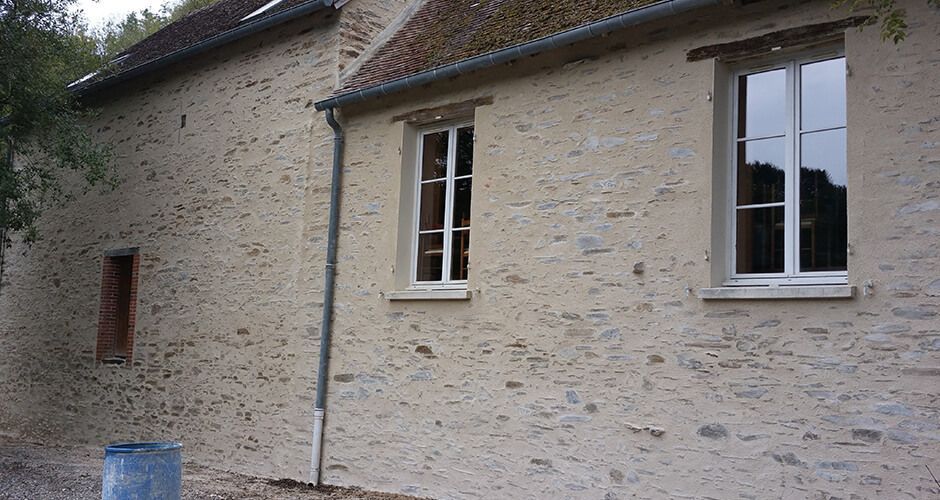 Un mur en pierre d'un bâtiment recouvert d'un enduit clair, avec deux fenêtres à cadre blanc et un petit volet en bois brun.