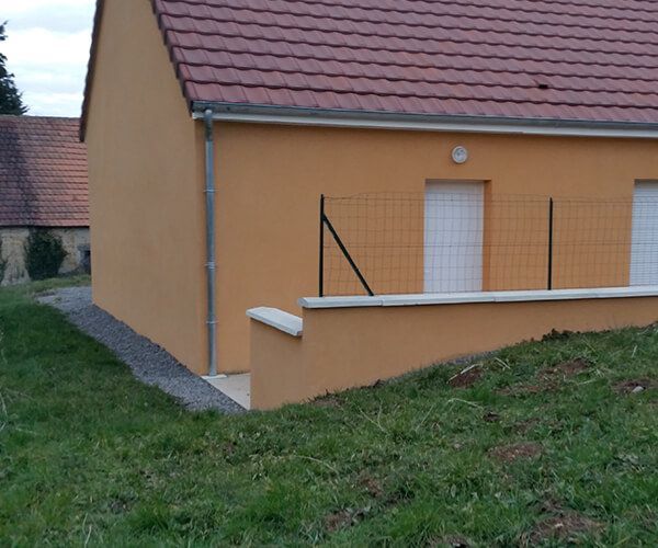 Une maison beige avec un toit de tuiles brun rougeâtre et une clôture métallique surmontant un muret périphérique bas, donnant sur une cour herbeuse.