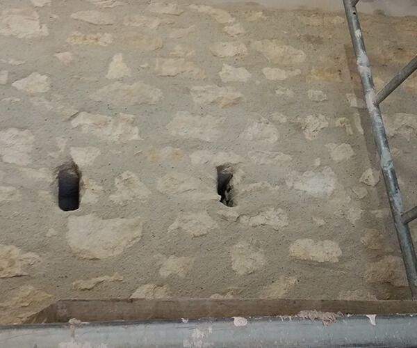 Un mur de pierre percé de deux petits trous rectangulaires sombres, vu du point de vue d'un échafaudage.