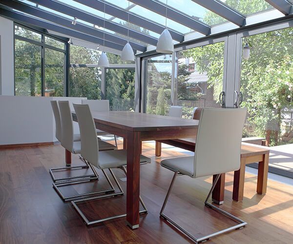 Une salle à manger avec une table en bois, plusieurs chaises grises modernes et une banquette sous un plafond de verre avec des suspensions lumineuses.
