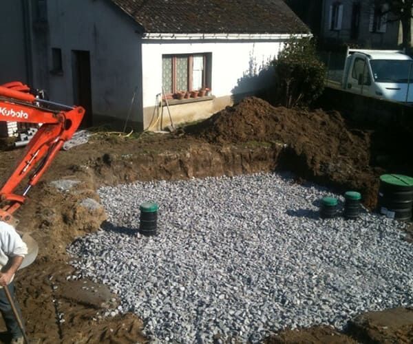 Une excavatrice est garée à côté d'une excavation dans un jardin résidentiel, remplie de gravier et comportant plusieurs tuyaux d'accès à la fosse septique de couleur verte.