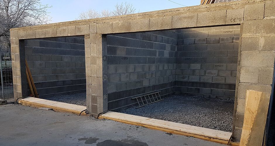 Un garage en blocs de béton à deux travées, en construction, avec des portes ouvertes et un sol intérieur en gravier.