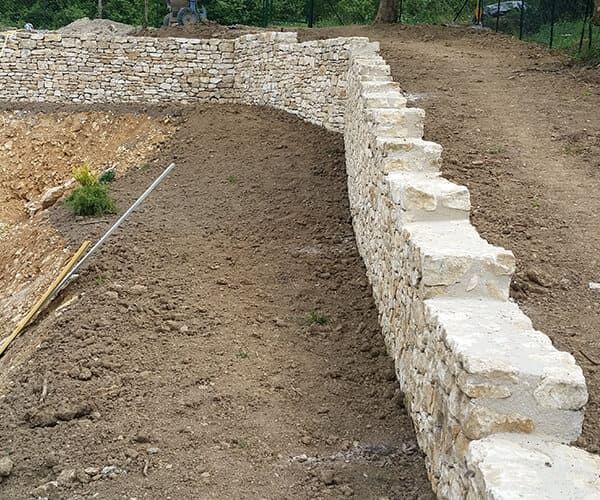 Un mur de soutènement en pierre longe un talus de terre dans un cadre extérieur, avec une petite plante verte à proximité.