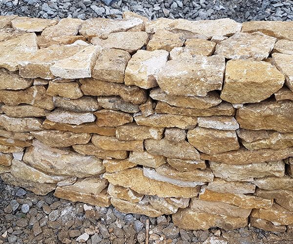Un muret construit en blocs de calcaire couleur miel de forme irrégulière, se détachant sur un fond de gravier sombre.