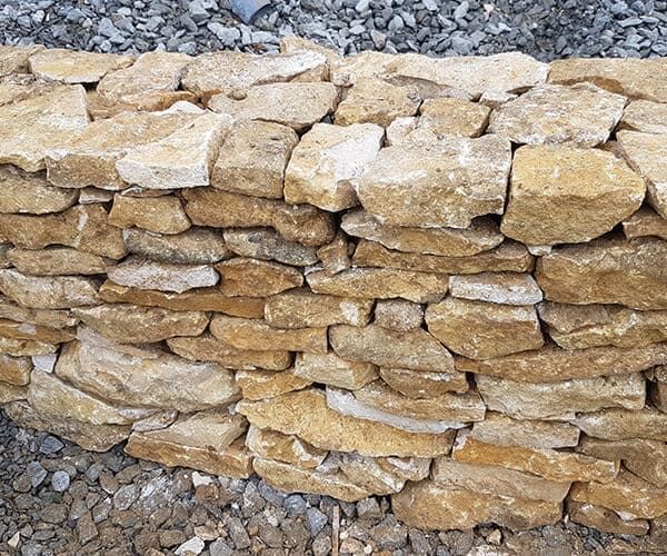Un mur en pierres sèches, construit à partir de roches rectangulaires brutes de couleur beige, se détachant sur un fond de gravier gris.