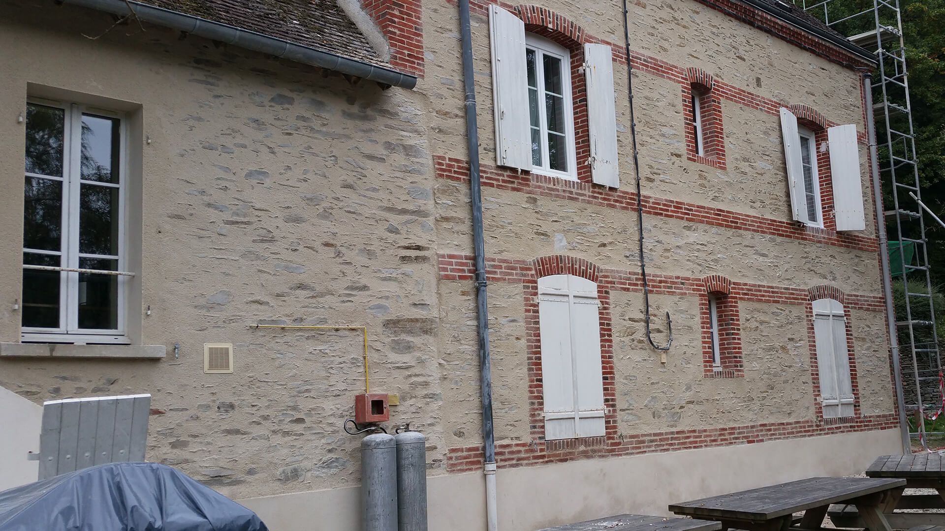 Un immeuble en pierre à plusieurs étages, avec des volets blancs, des accents de briques rouges et un mur en plâtre uni sur la gauche.