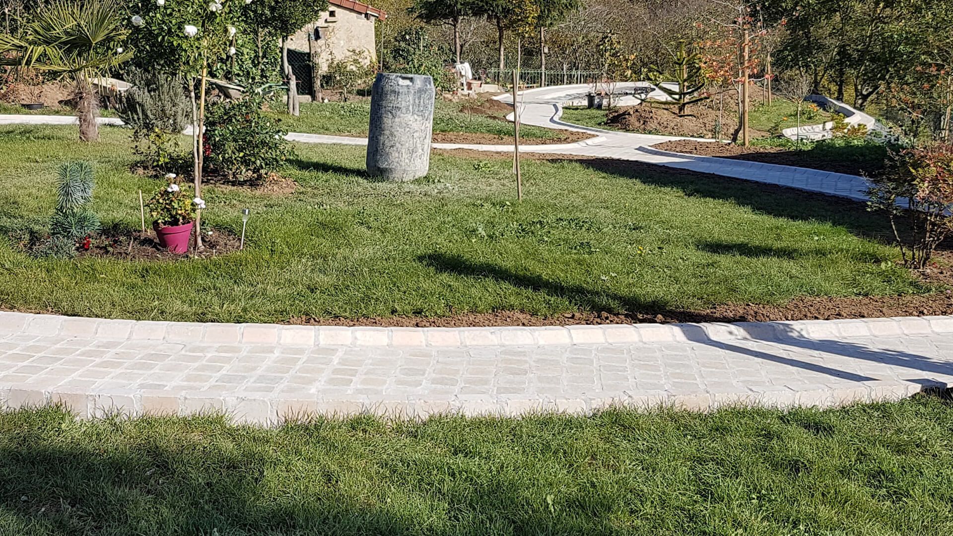 Un chemin de pierres claires serpente à travers un jardin herbeux arboré, orné d'un pilier de pierre grise.