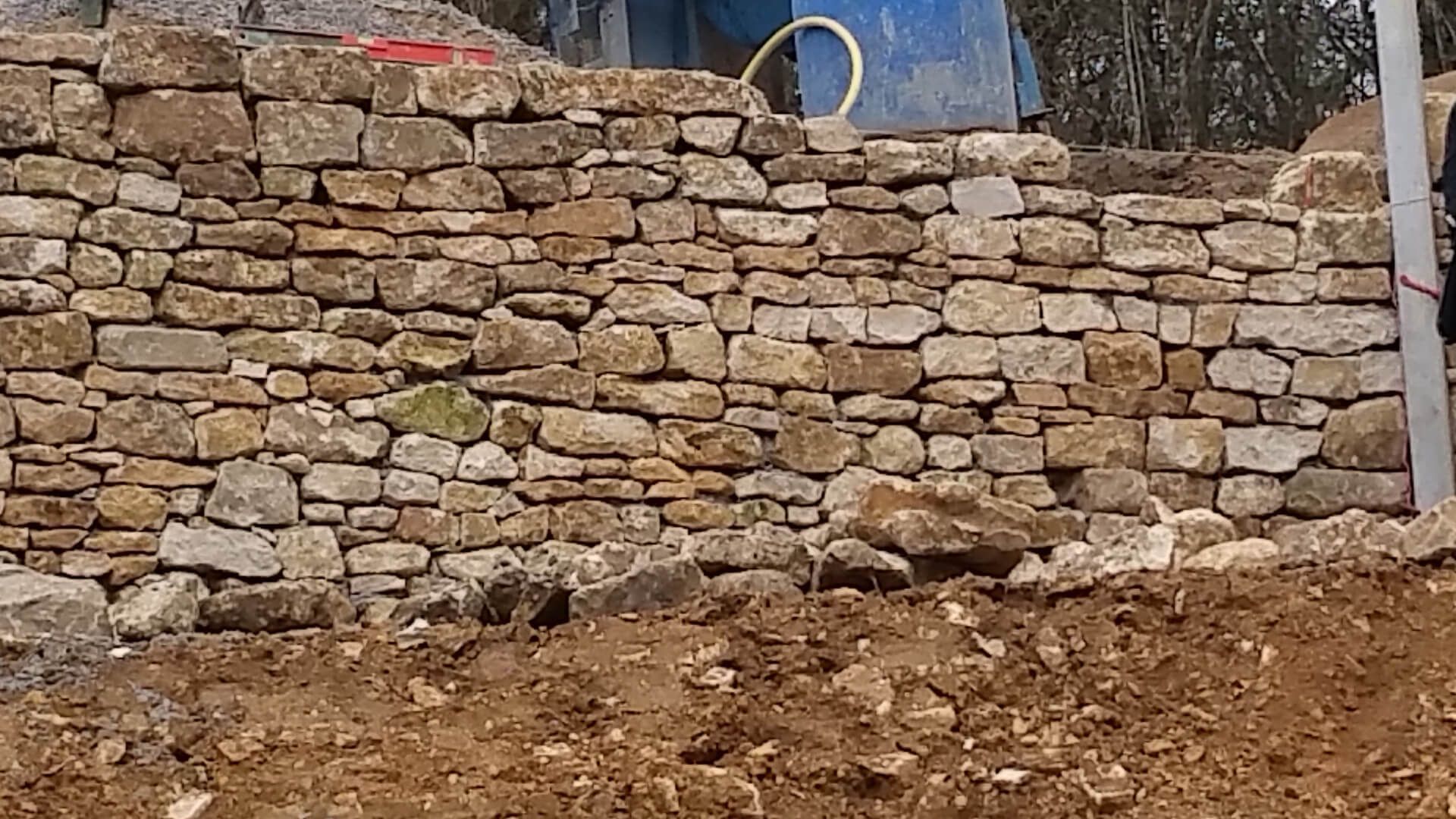 Un muret en pierres sèches se dresse derrière un monticule de terre excavée, avec un poteau métallique vertical à droite et une bâche bleue au-dessus.