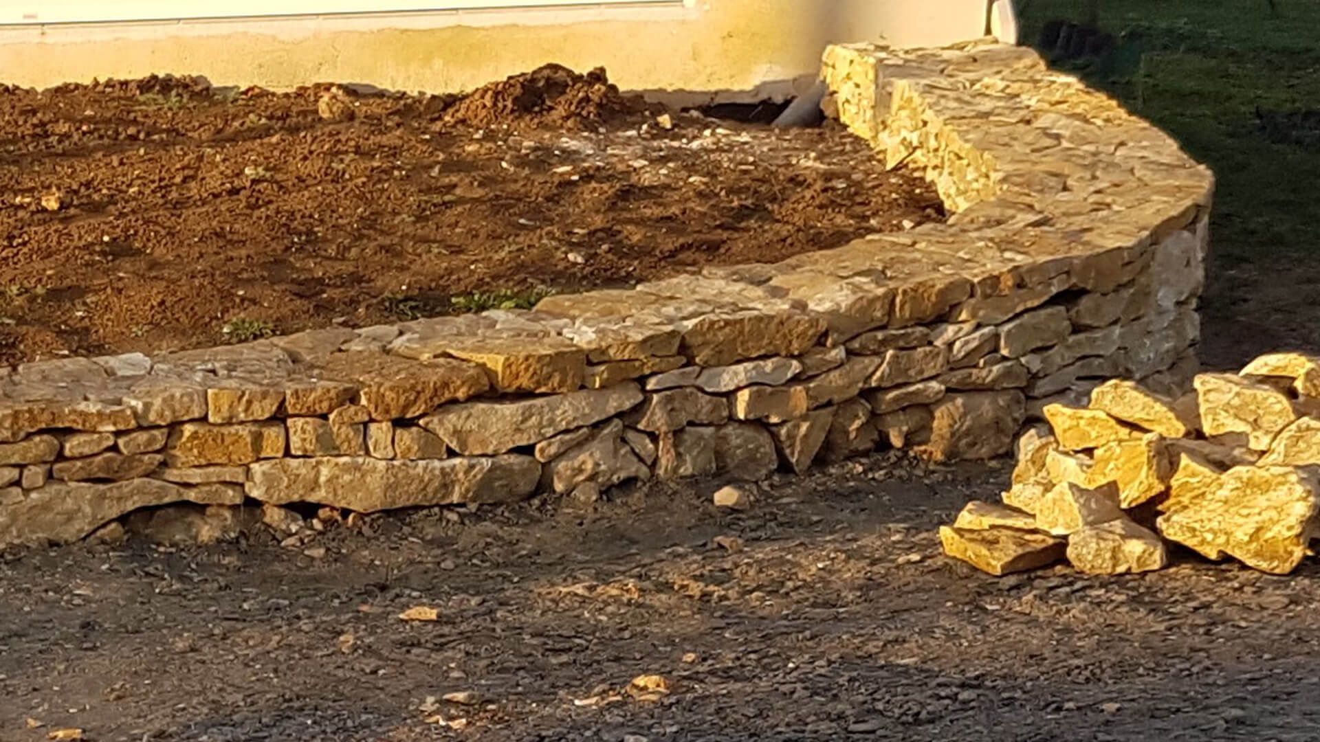 Un muret bas et incurvé en pierres sèches beiges et brutes borde un lit de terre sombre à côté d'un bâtiment de couleur claire.