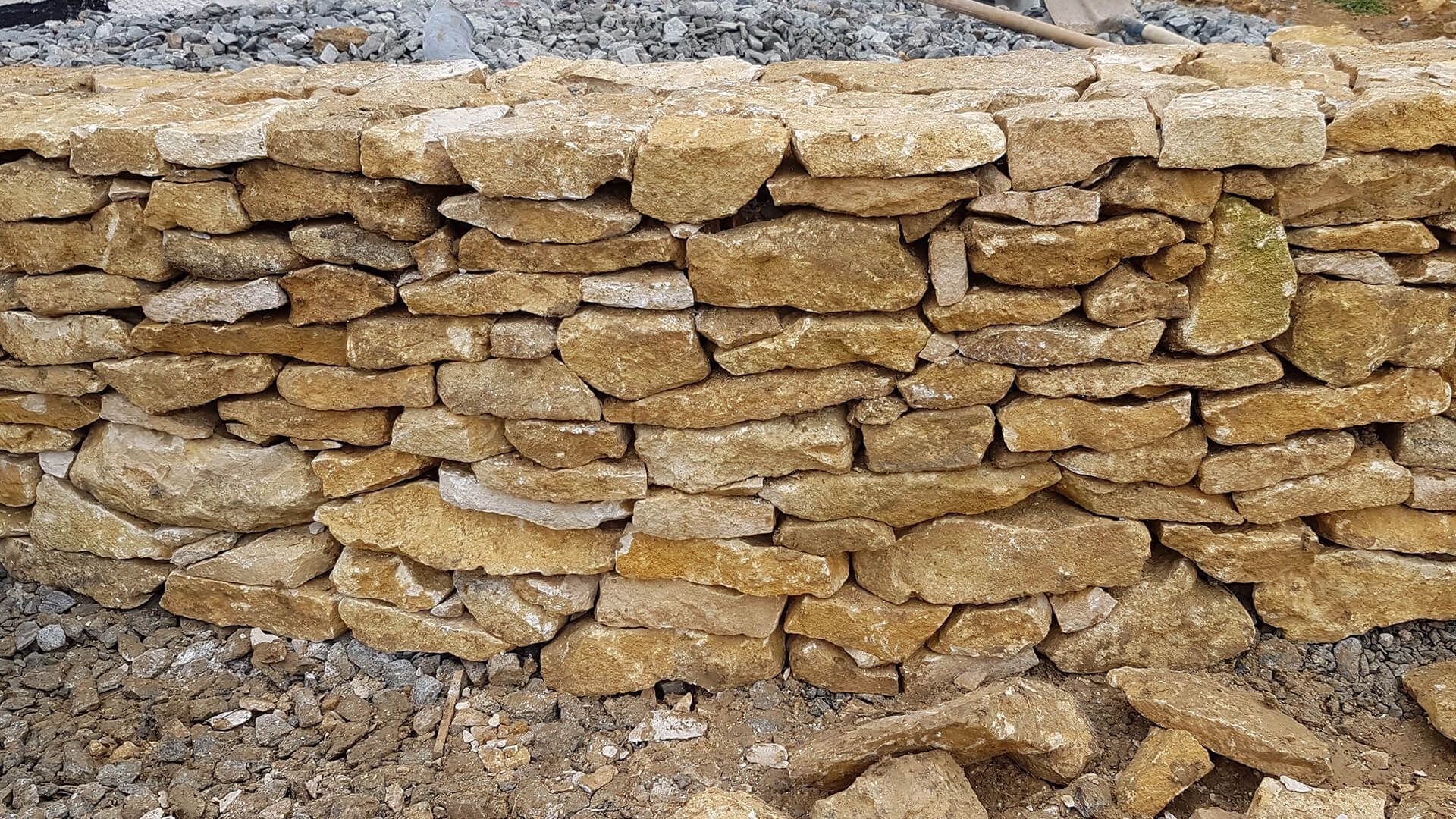 Un mur en pierres sèches, composé de pierres rectangulaires brutes, brun doré, se détachant sur un fond de gravier épars.