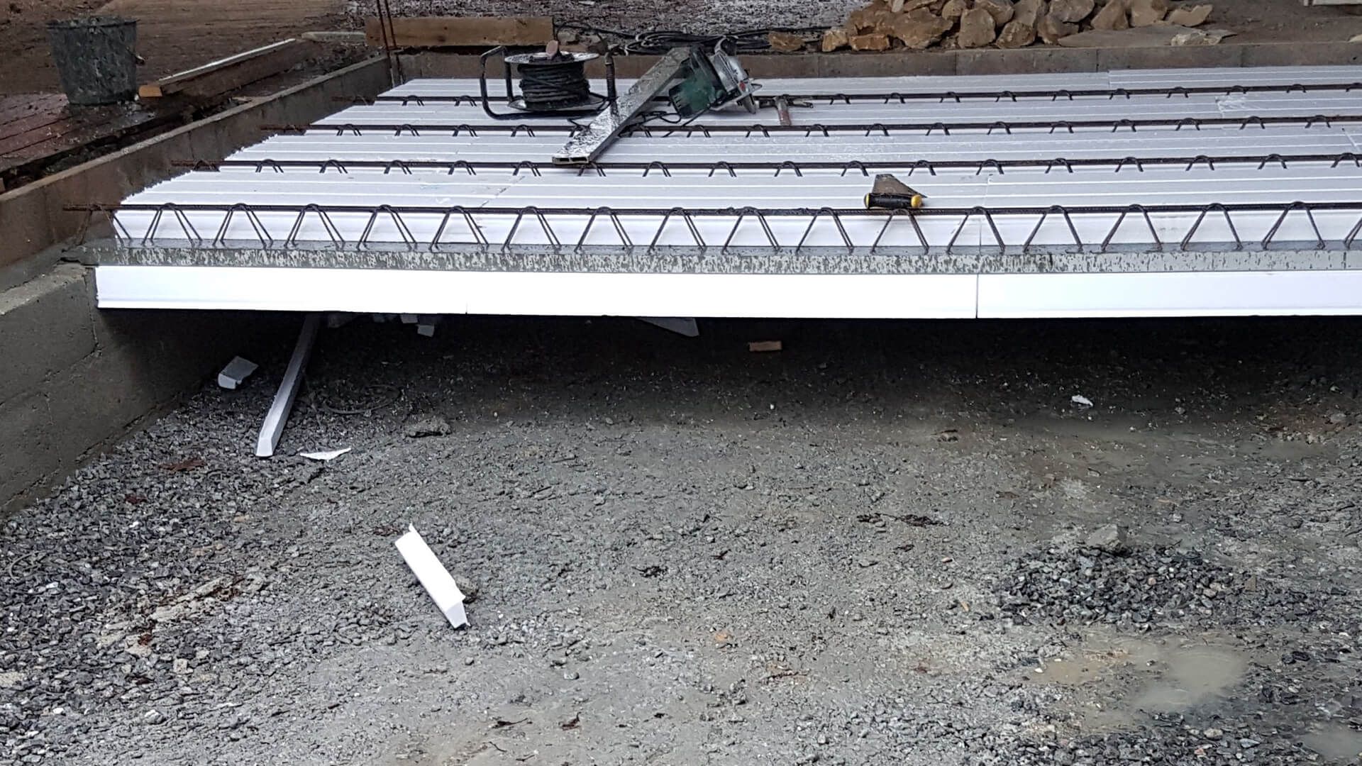 Des fermes en acier d'armature reposent sur des panneaux d'isolation en mousse blanche, eux-mêmes posés sur un chantier de construction recouvert de gravier.
