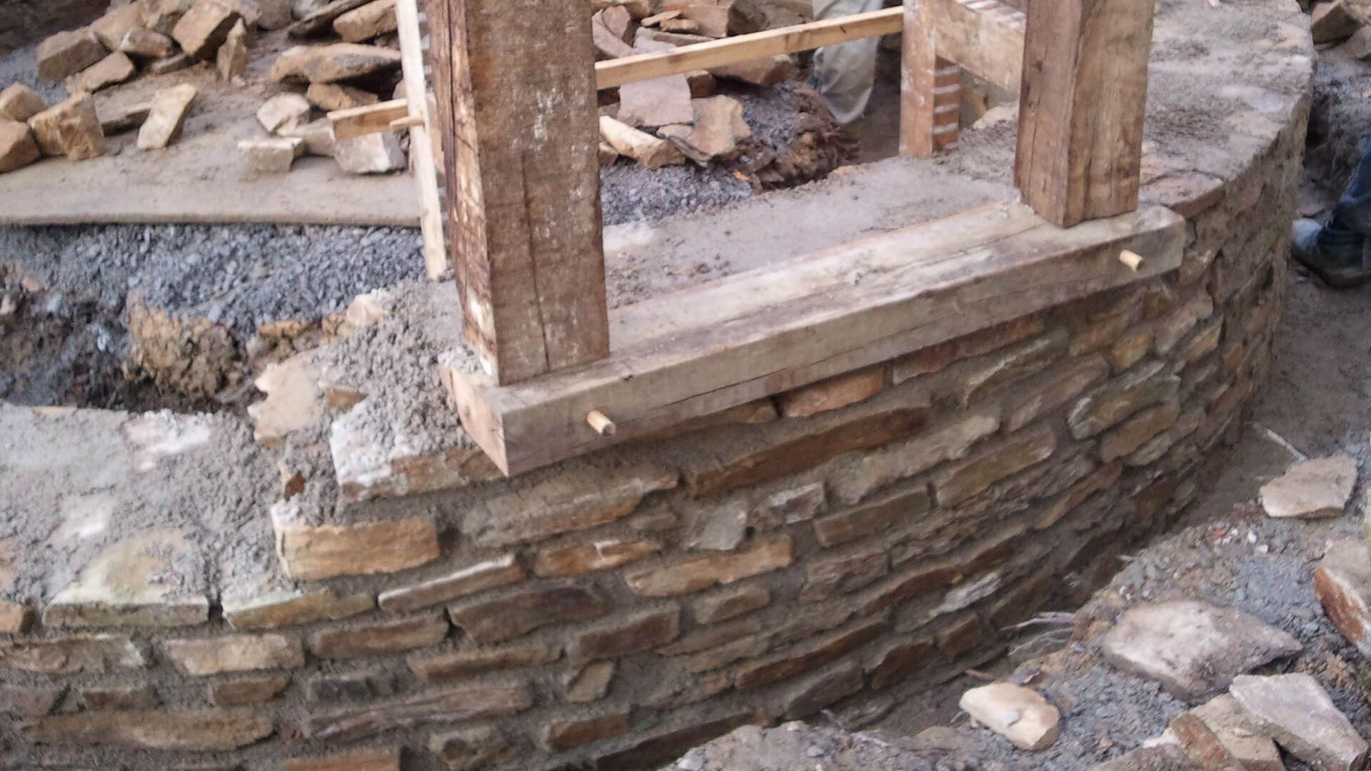 Un mur de fondation incurvé en pierre est en construction, avec des poteaux à ossature bois mis en place avec du mortier.