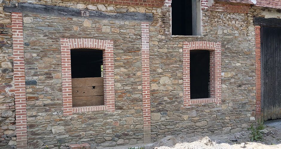 Un mur en pierre patinée par le temps, avec deux fenêtres rectangulaires encadrées de briques et une porte en bois sur la droite.