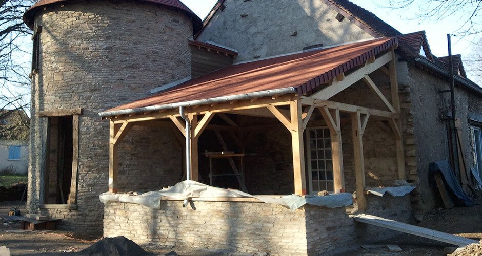 Rénovation extérieure d'un bâtiment en pierre avec un porche à ossature bois nouvellement construit et un toit en tuiles de terre cuite.