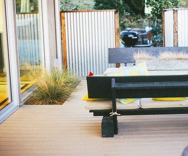 Une terrasse avec un plancher en bois, un banc noir avec des coussins jaunes, un petit carré de gazon et des écrans en tôle ondulée.