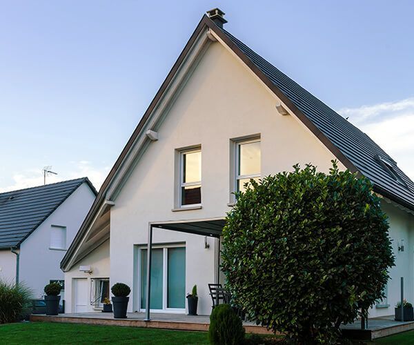 Maison blanche à pignon avec patio, sièges extérieurs et un grand arbuste rond dans une cour herbeuse sous un ciel bleu.
