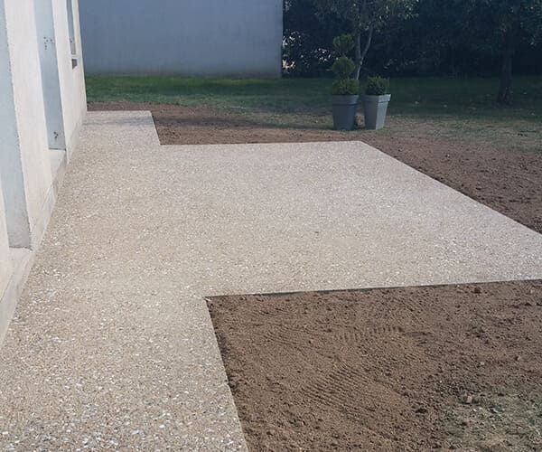 Une terrasse en gravier clair s'étend du côté d'une maison moderne jusqu'à un jardin parsemé de zones de terre nue.