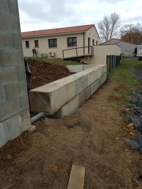 Chantier de construction extérieur avec mur de soutènement en béton, monticule de terre et maison beige en arrière-plan