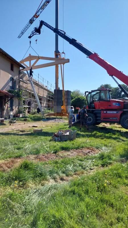 Une grue soulève une charpente en bois sur un chantier de construction, à côté d'un camion rouge sur un terrain herbeux.