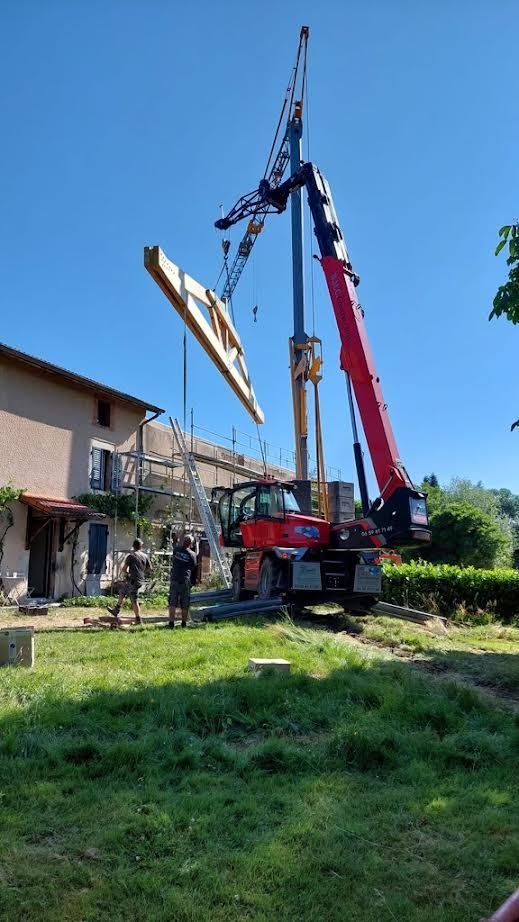 Une grue soulève une grande charpente à côté d'une maison dans une cour herbeuse sous un ciel bleu dégagé.