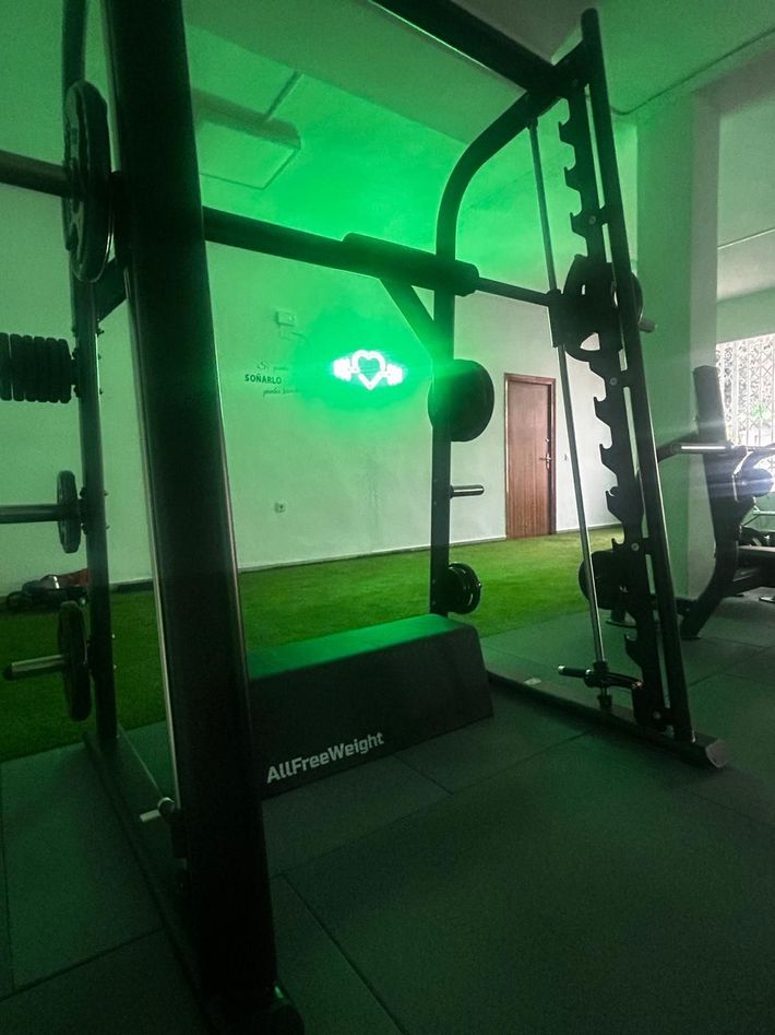 Banco de gimnasio con mancuernas y discos de pesas frente a un espejo y ventanas.