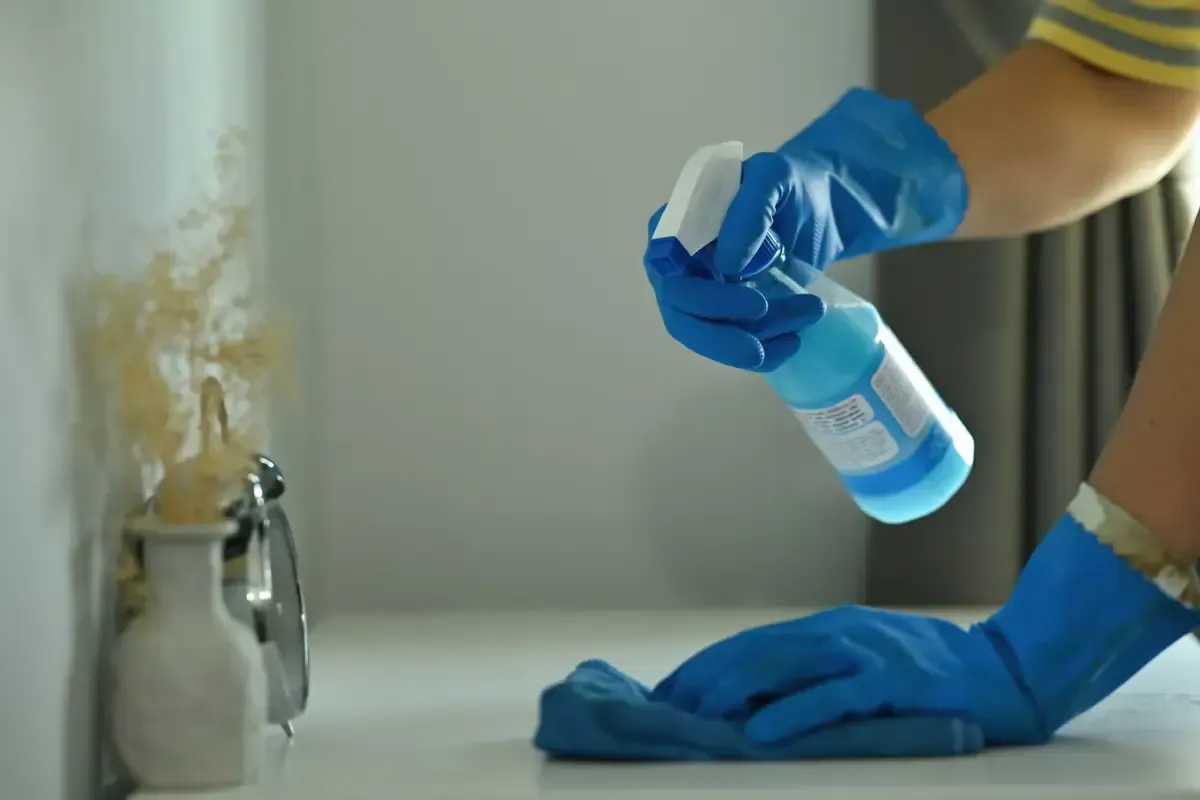 Persona con guantes azules, limpiando una superficie blanca con un atomizador y un paño.