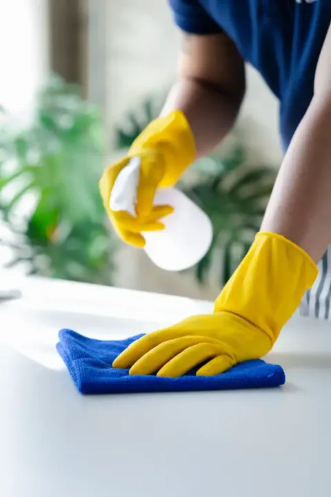 Una persona que lleva guantes amarillos rocía y limpia una superficie blanca con un paño azul.