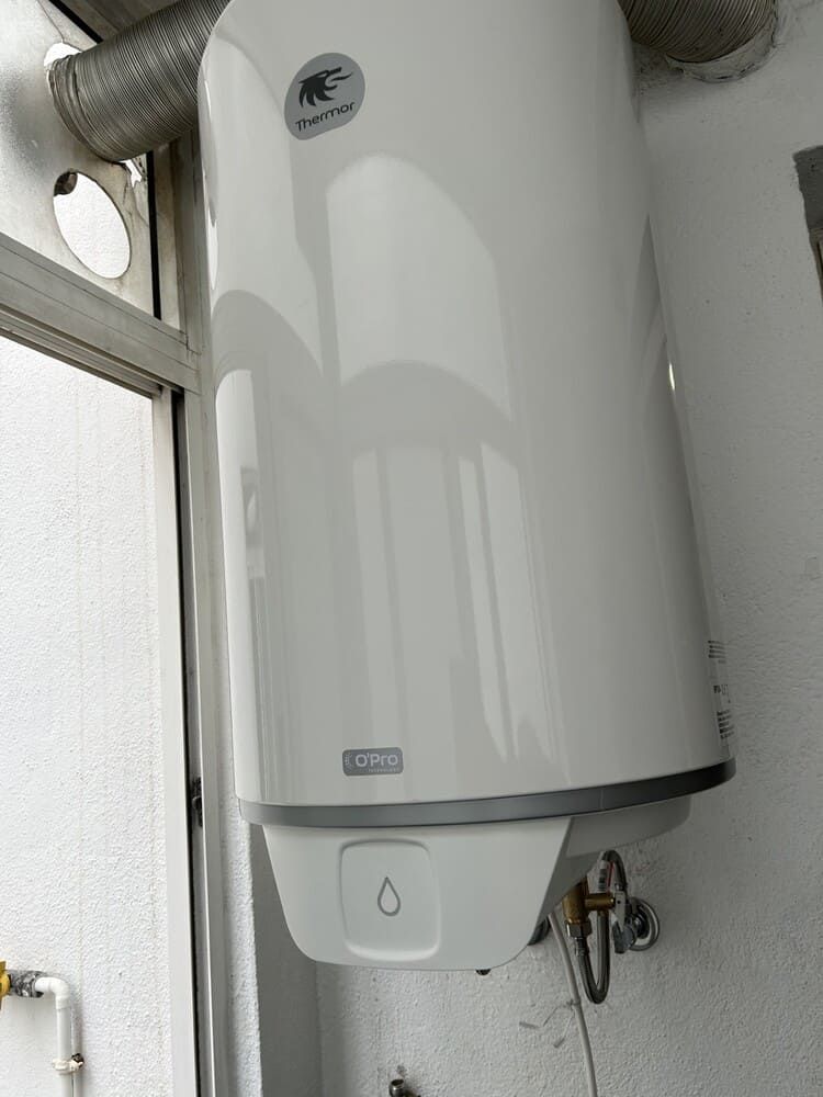 Calentador de agua blanco montado en la pared, ubicado en un pequeño cuarto de servicio, con tuberías y una válvula de drenaje debajo.