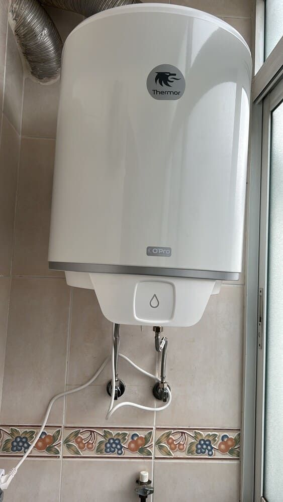Calentador de agua blanco montado en la pared de un baño, con tuberías y revestimiento de azulejos debajo.