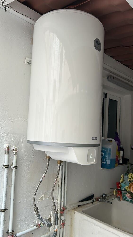 Calentador de agua blanco montado en la pared sobre un fregadero, con tuberías expuestas en un área de servicio.