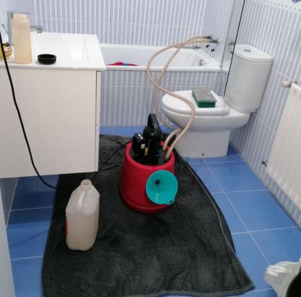 Baño con cubo de fregar sobre alfombra negra, jarra blanca, lavabo con mueble, inodoro y radiador sobre suelo de baldosas azules.