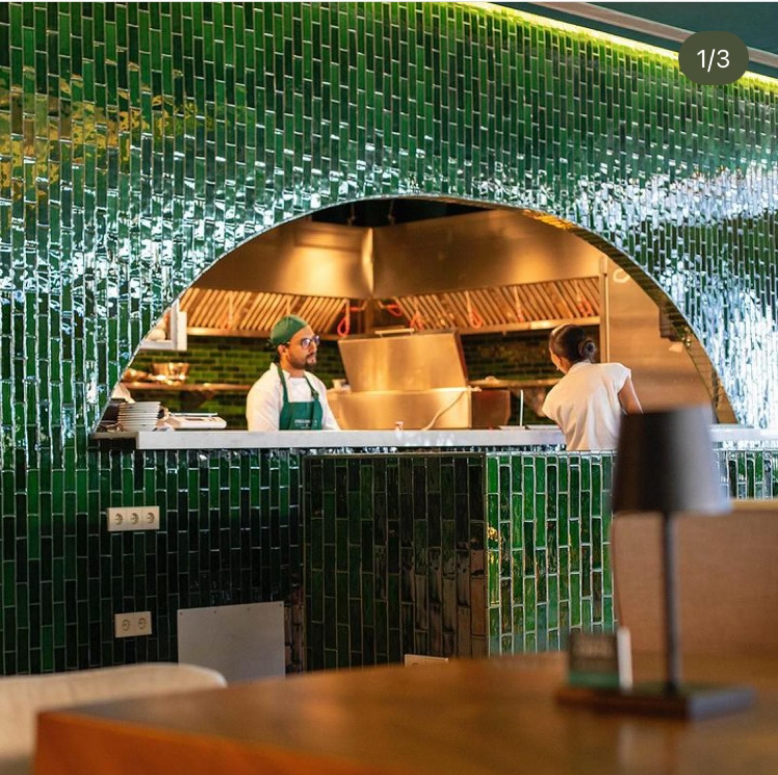 Una cocina de restaurante de paso, enmarcada con azulejos verdes. Dos cocineros trabajan en el interior.