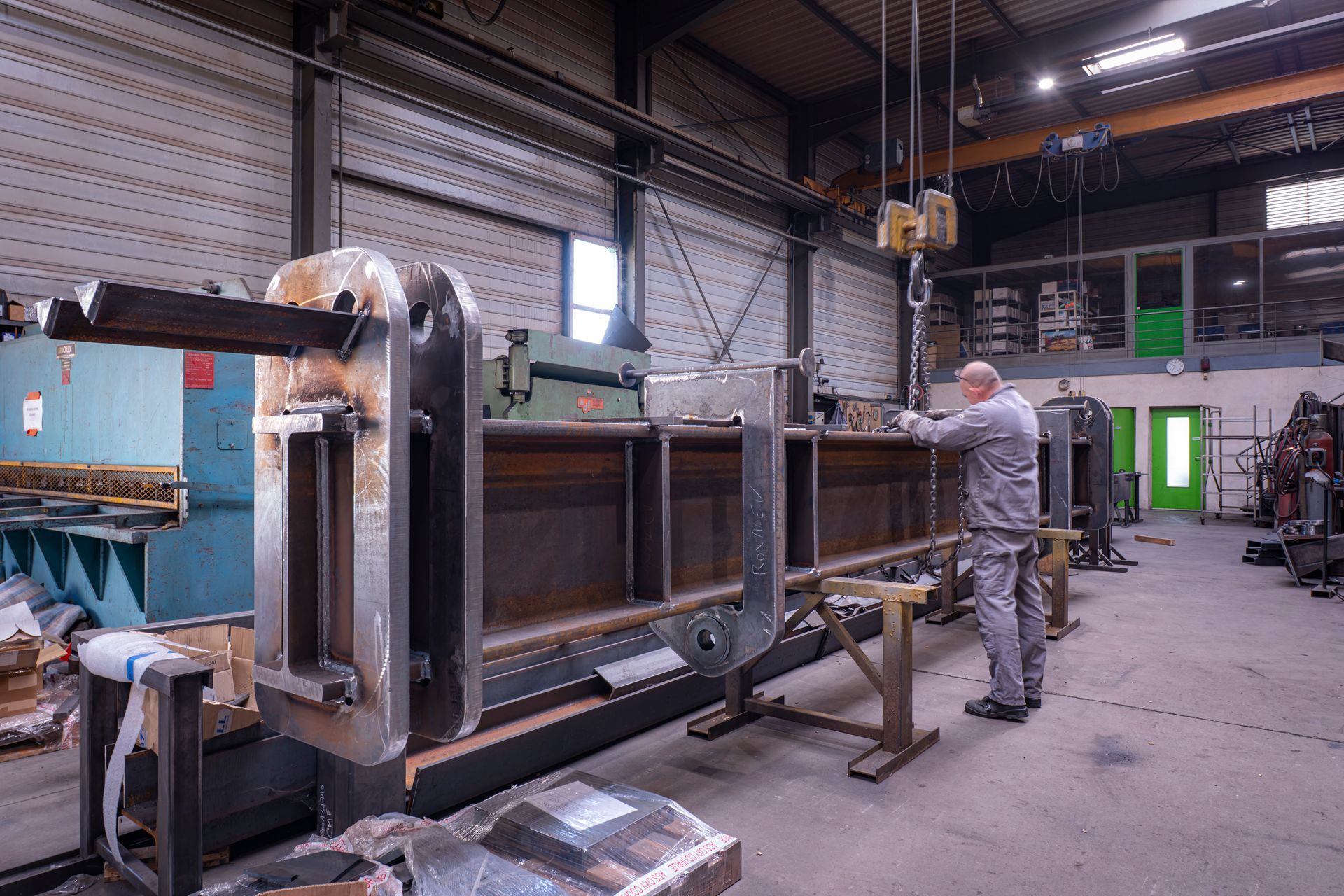 Un homme travaille dans un atelier de métallurgie sur une grande structure en acier. On aperçoit un pont roulant et des machines.