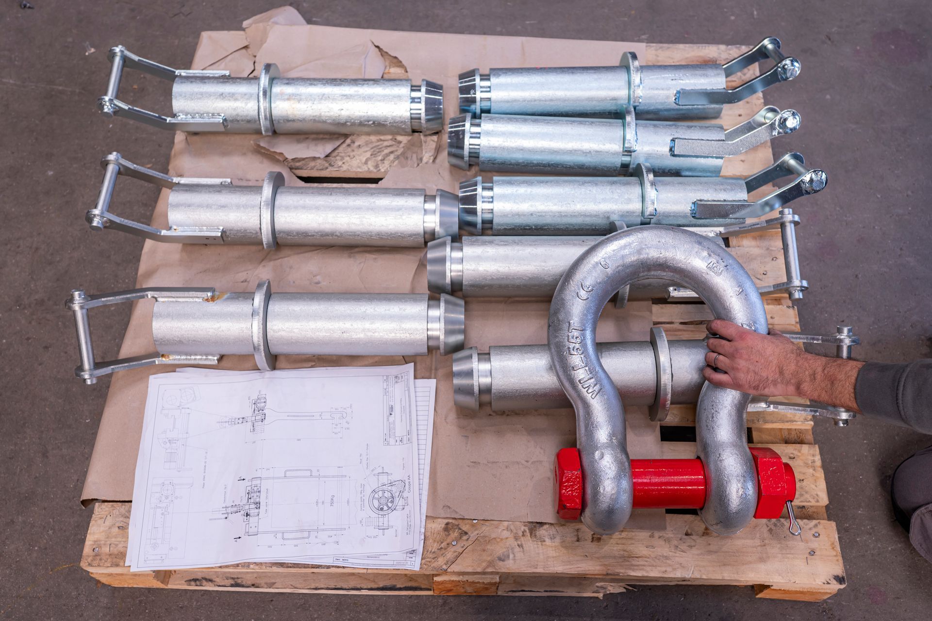 Palette contenant des pièces industrielles métalliques ; une main tient une grande manille rouge à côté de plans.