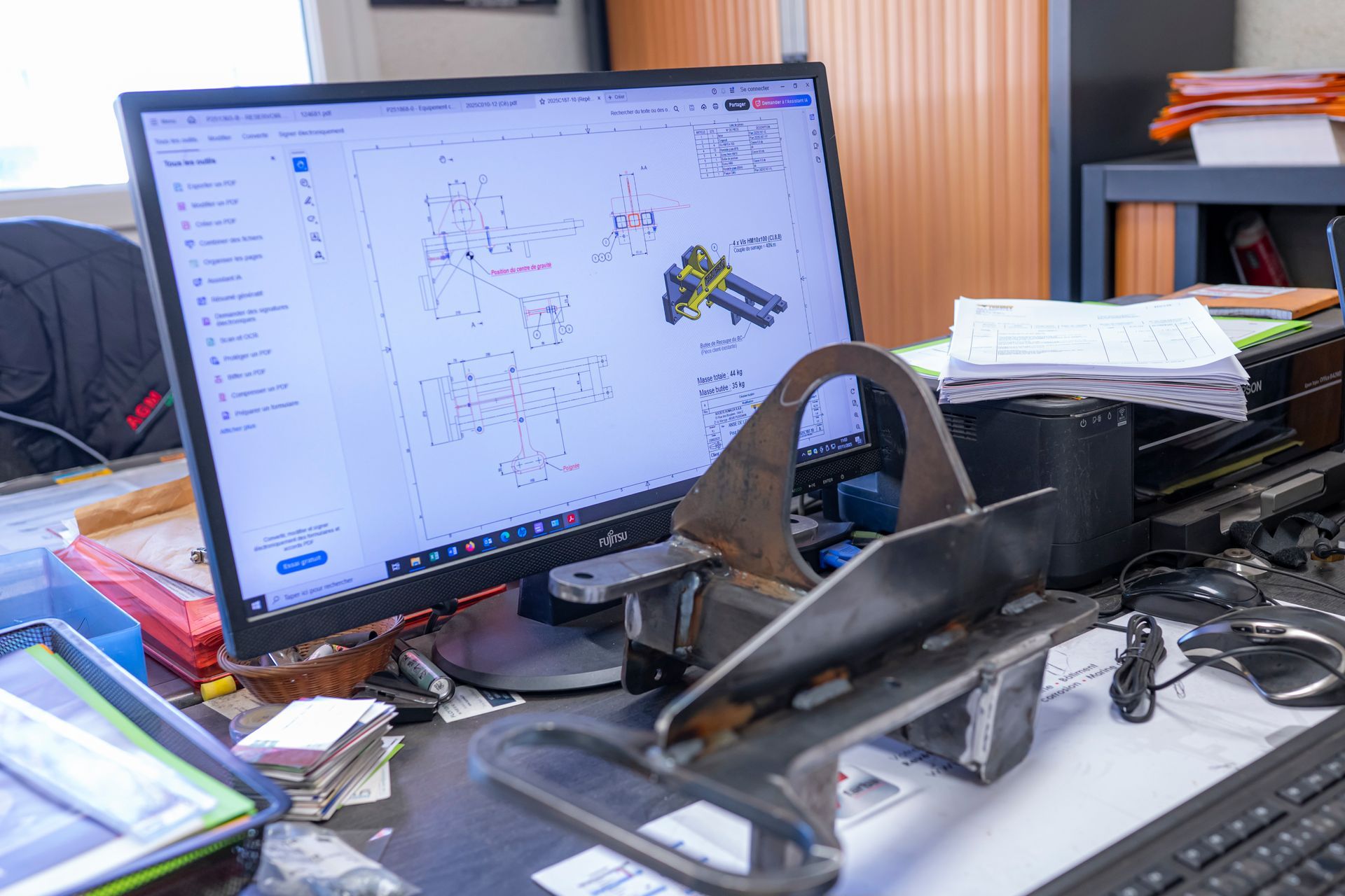 Un écran d'ordinateur affiche un dessin 3D, à côté d'une pièce métallique posée sur un bureau encombré dans un bureau.