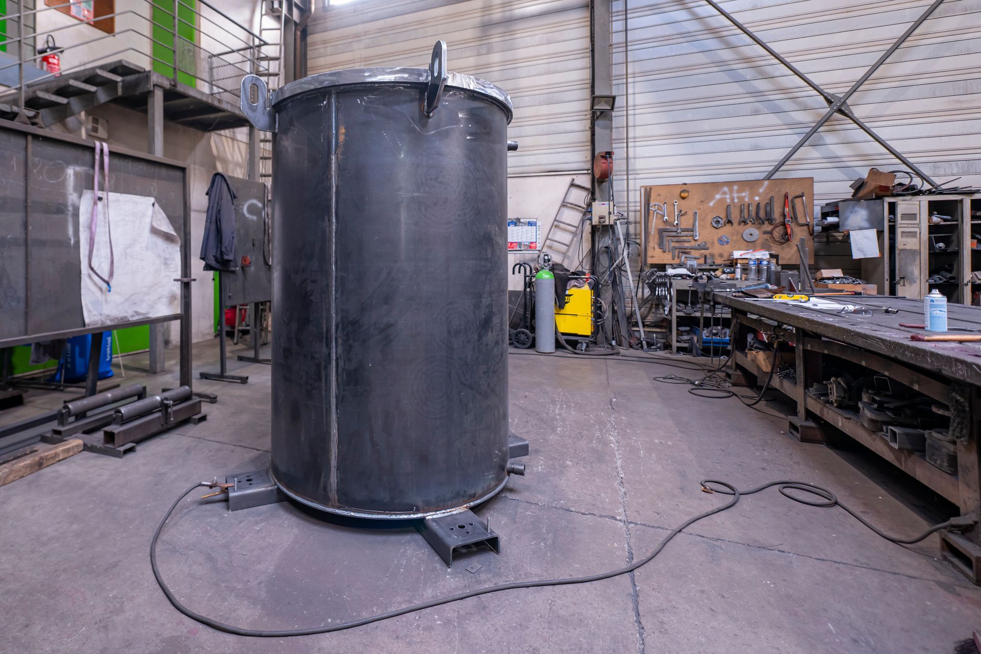 Grand réservoir cylindrique noir dans un atelier ; outils et équipements de soudage en arrière-plan.