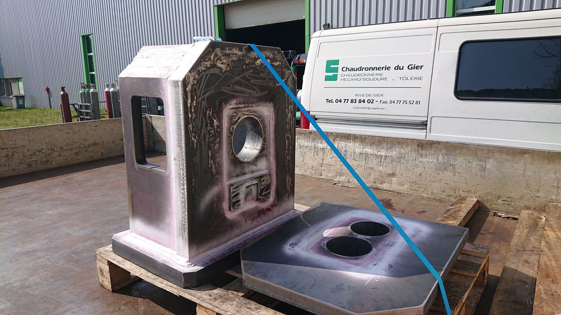 Pièces industrielles métalliques soudées sur une palette, probablement destinées à une machine, à l'extérieur d'un bâtiment.