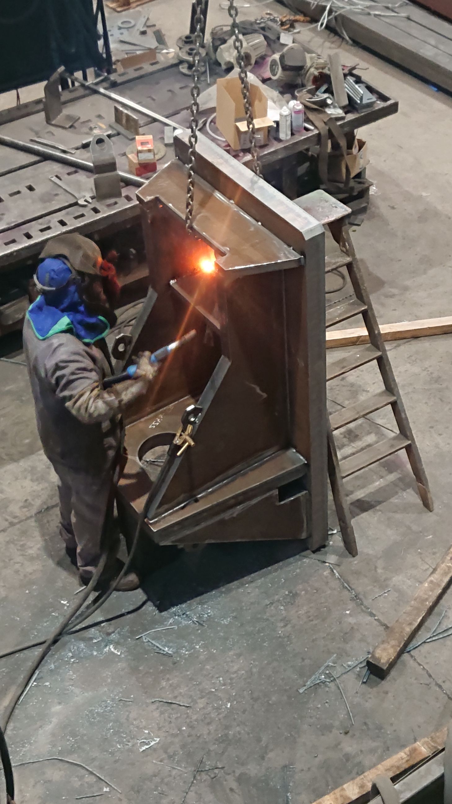 Un soudeur en tenue de protection travaille sur une grande structure métallique dans un atelier.