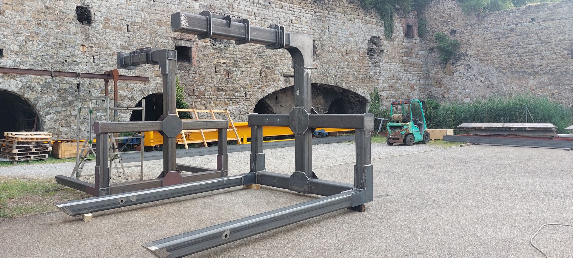 Gros accessoire métallique industriel pour chariot élévateur, installé à l'extérieur devant un mur en pierre.