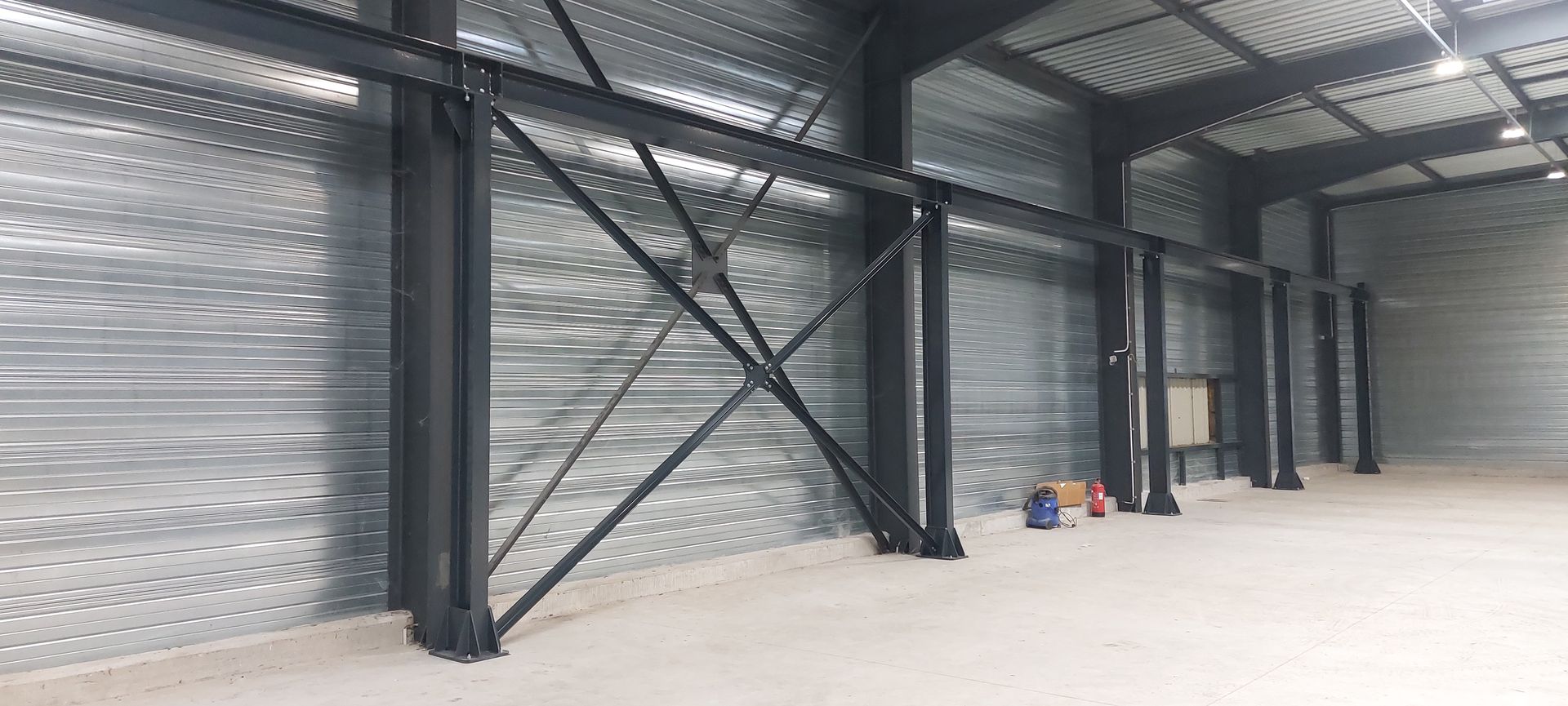 Intérieur d'un grand bâtiment métallique vide, avec des poutres structurelles et un sol en béton.