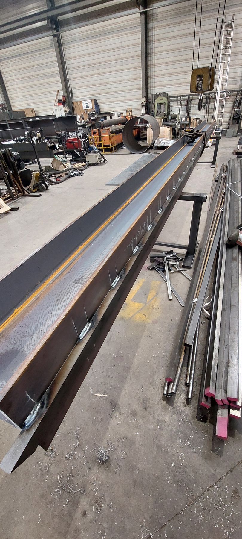 Une longue poutre métallique dans un atelier, avec des outils et du matériel en arrière-plan.