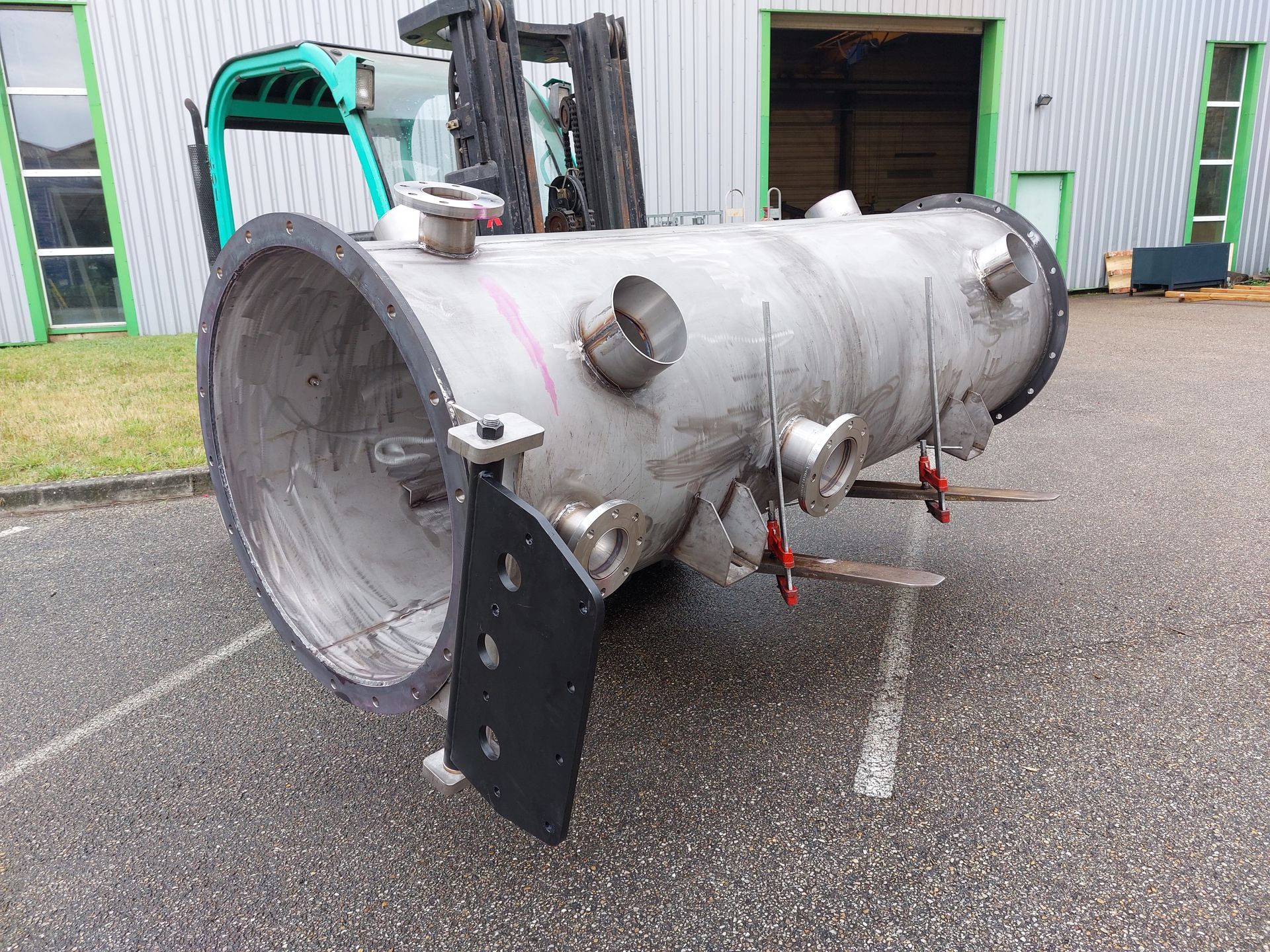 Un grand réservoir industriel cylindrique en acier inoxydable est déplacé par un chariot élévateur sur une surface asphaltée.