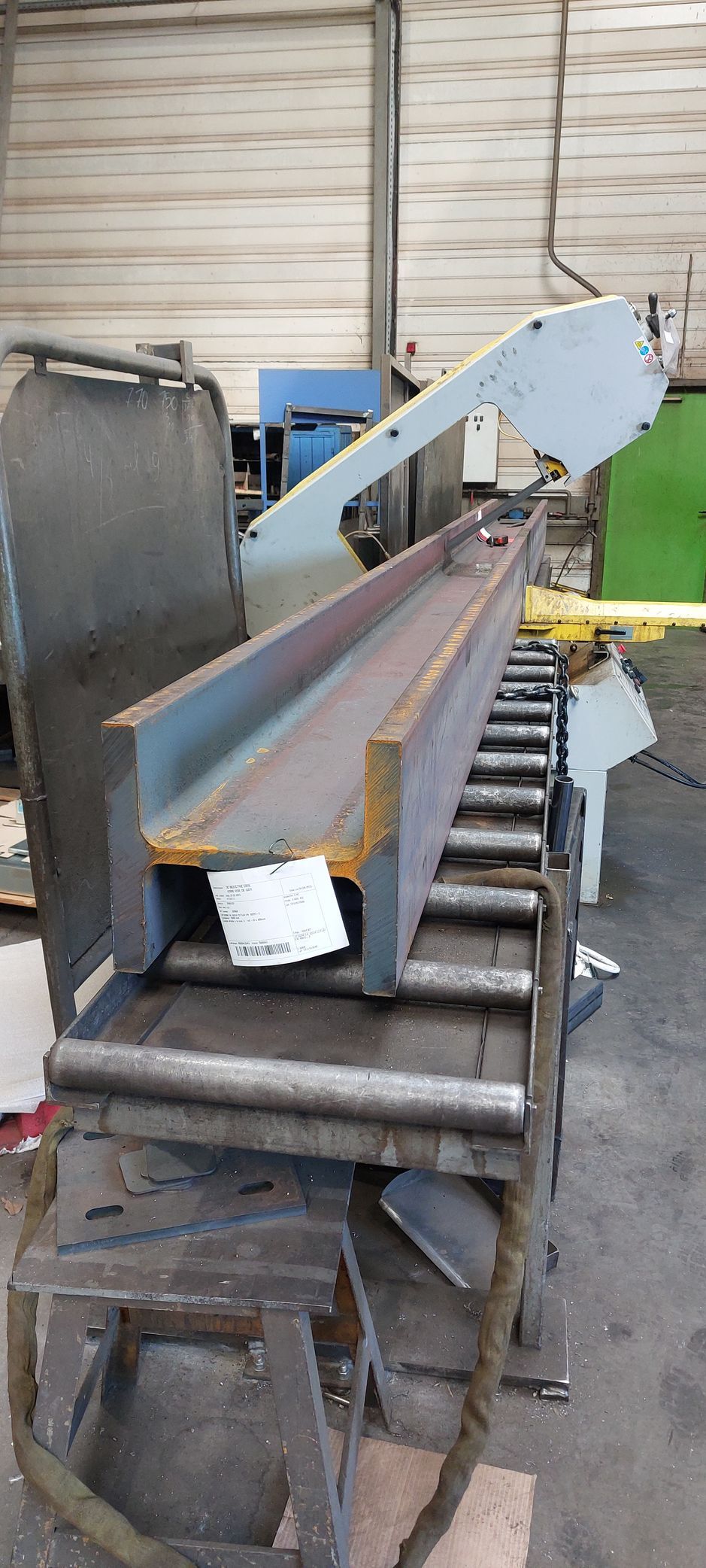 Une longue poutre d'acier sombre sur un convoyeur à rouleaux métalliques, prête à être coupée par une scie à ruban.