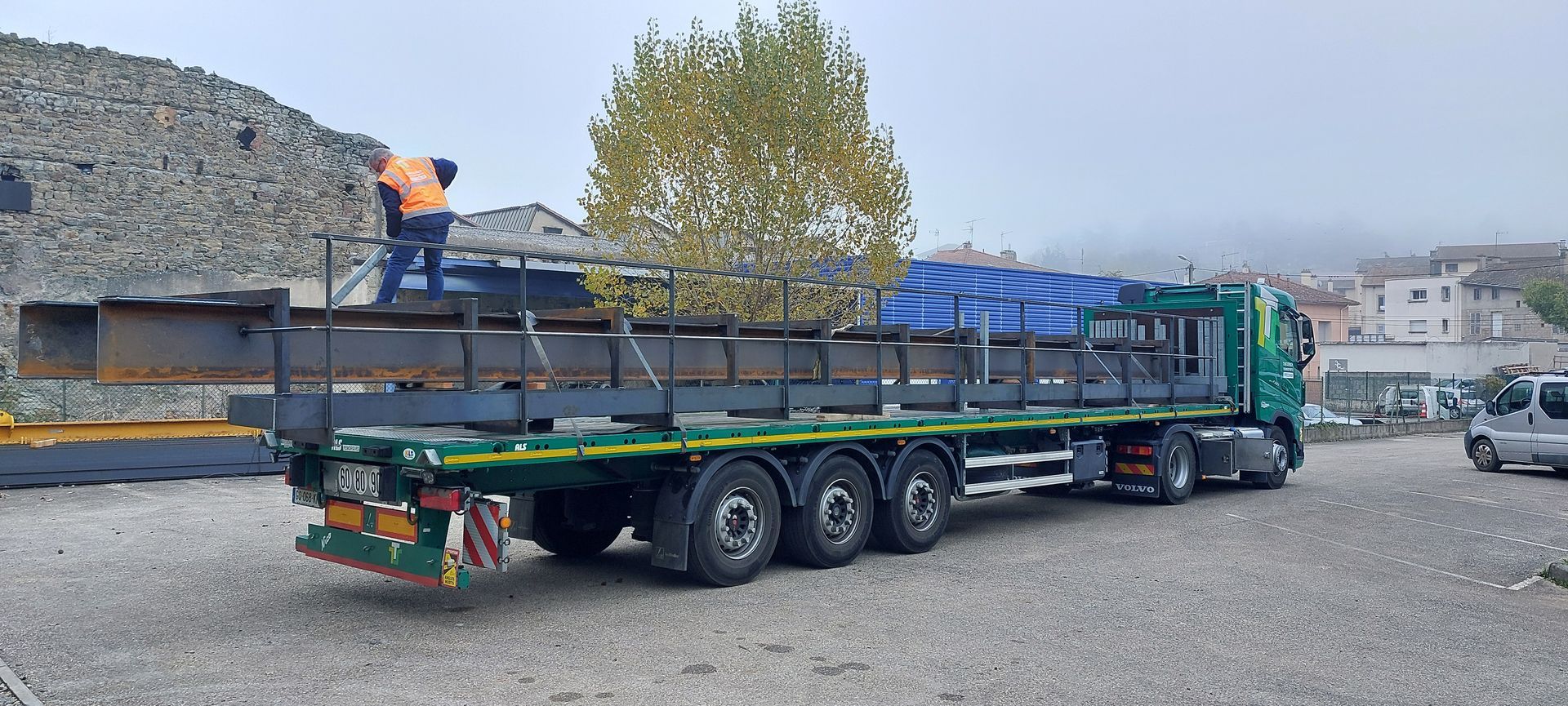 Camion transportant des poutres d'acier, un ouvrier en gilet orange, décor extérieur avec un bâtiment et une zone de gravier.