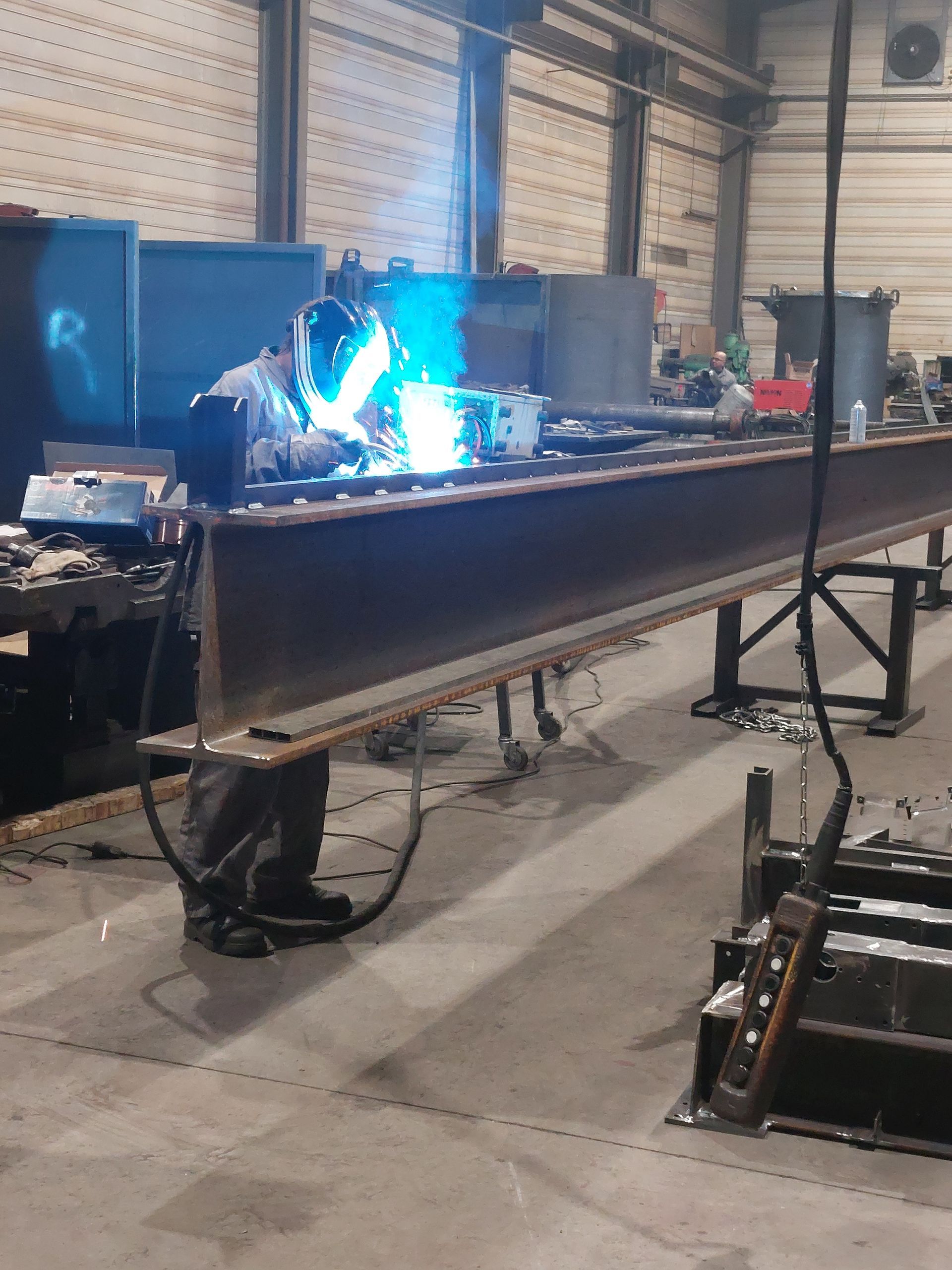 Un soudeur portant un équipement de protection soude une poutre métallique dans un atelier. Arc bleu vif.
