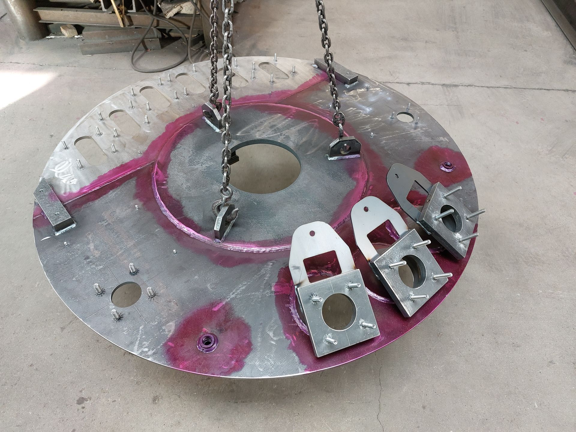 Grand disque métallique avec supports soudés, suspendu par des chaînes. Les soudures sont mises en valeur par une teinture rose ; décor d'atelier.