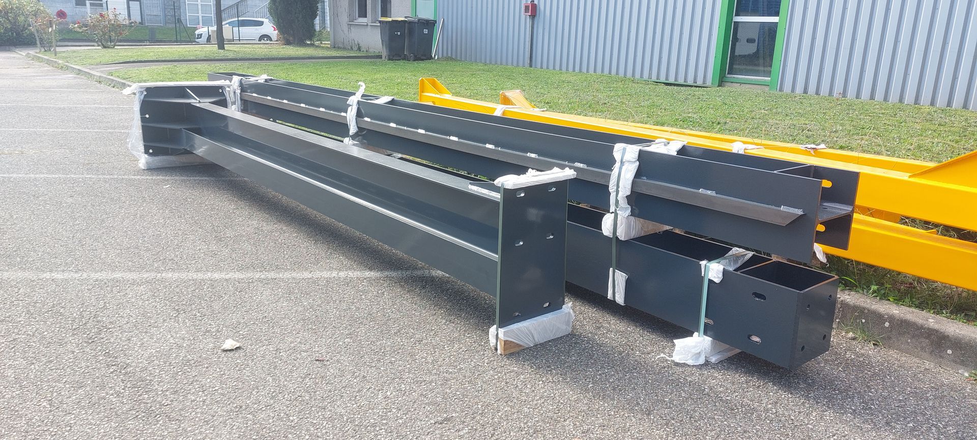 Des poutres métalliques gisent sur l'asphalte à côté d'un bâtiment. Des structures jaunes et vertes sont visibles en arrière-plan.